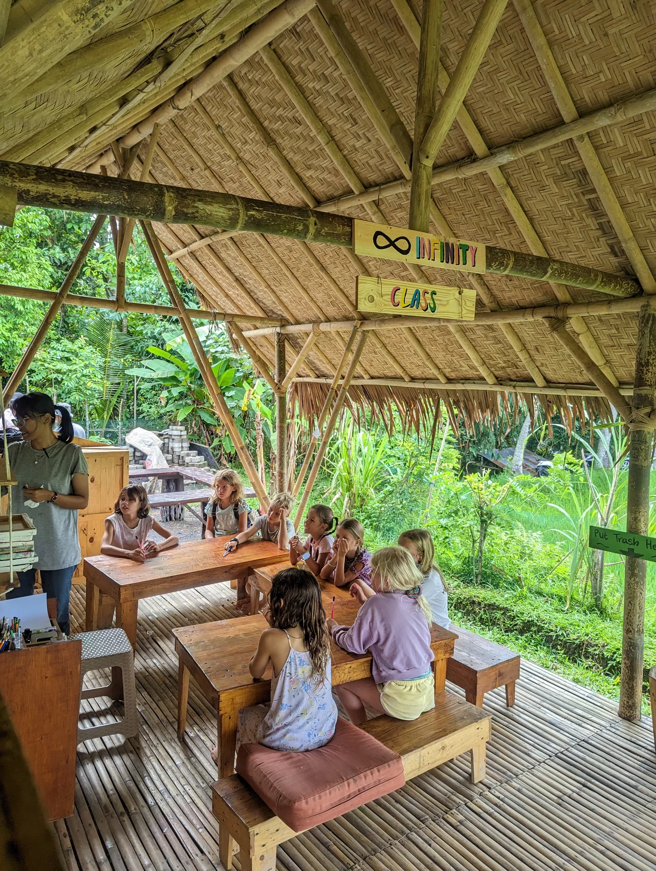 A teacher and children learning at wooden tables in an open bamboo classroom surrounded by greenery.