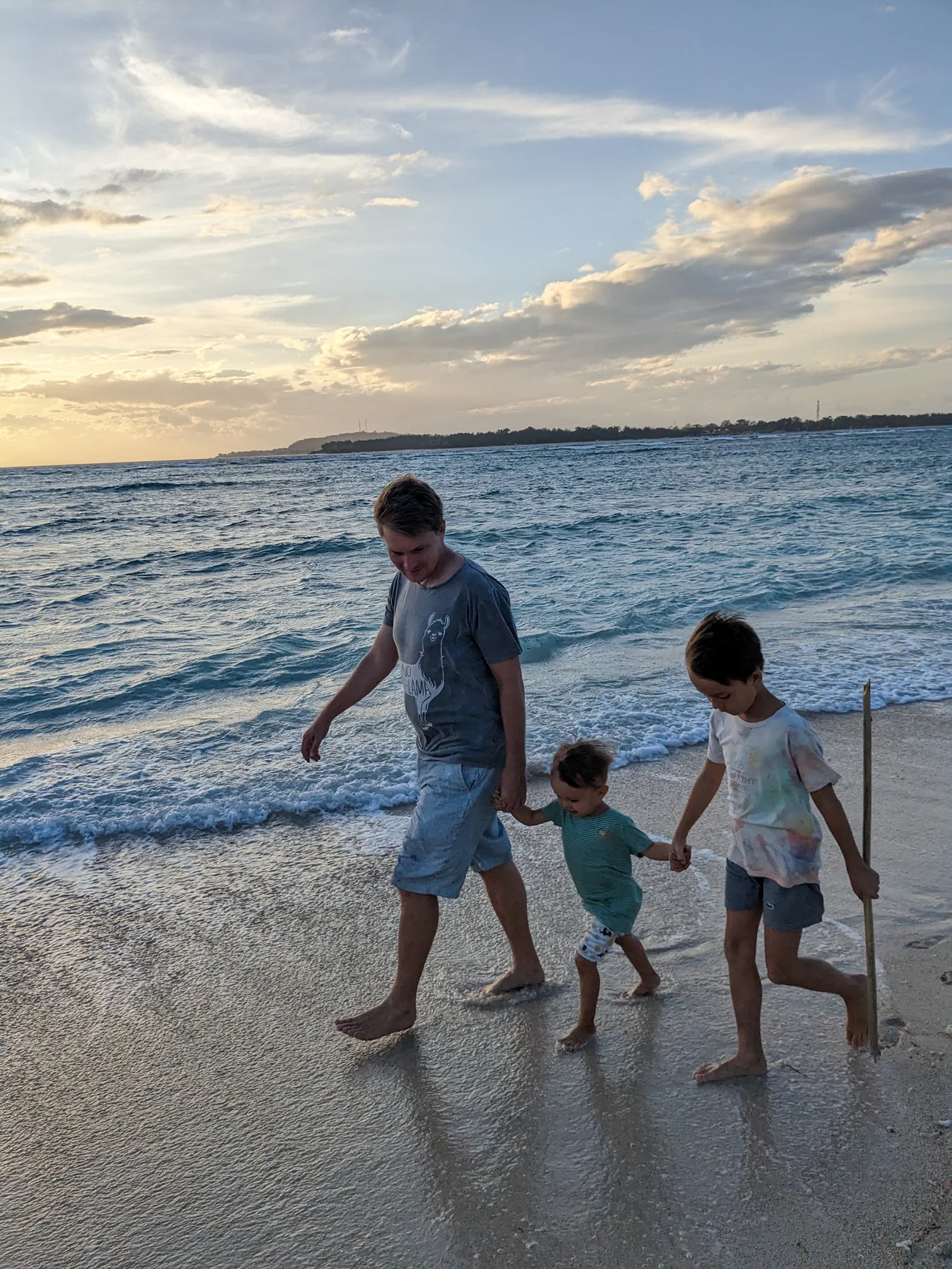 A father and two children walking hand in hand along the water's edge at sunset.
