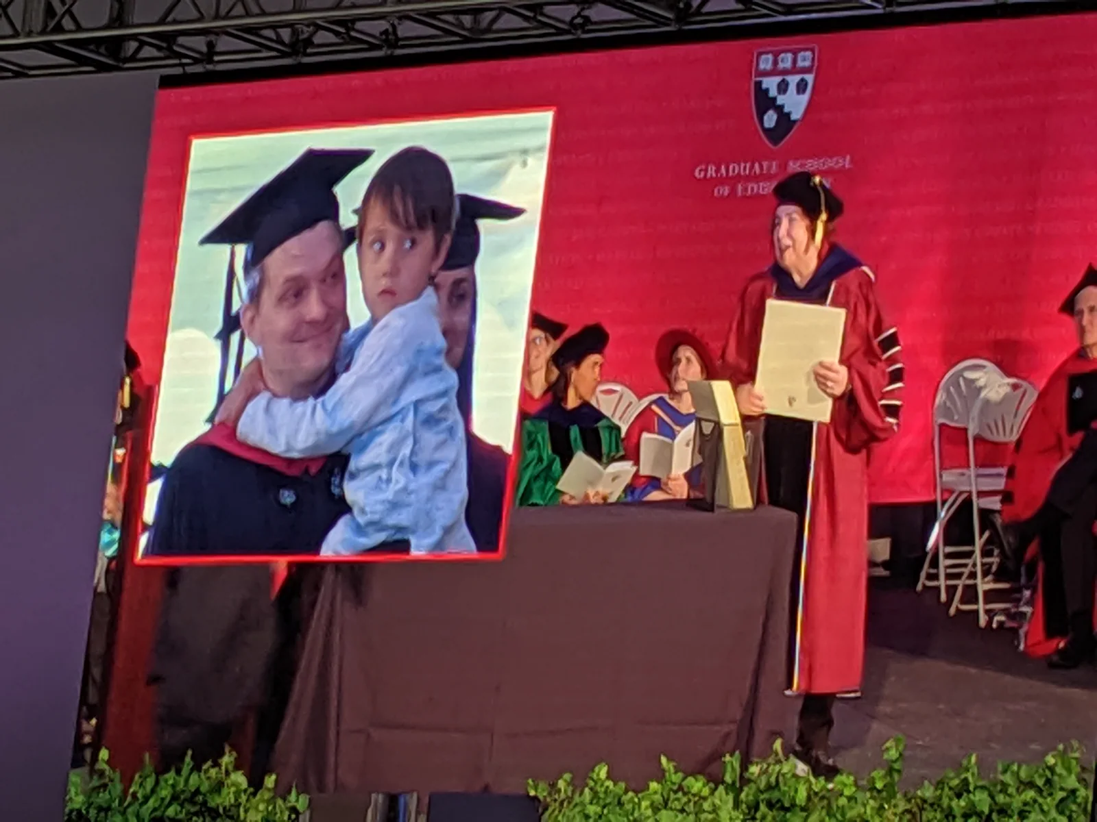 Harvard Graduate School of Education graduation ceremony, with a graduate holding a child shown on the large screen.